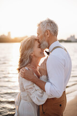 Stylish couple of elderly newlyweds stand embracing on river bank. Man kisses woman on forehead.