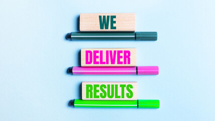On a light blue background, there are three multi-colored felt-tip pens and wooden blocks with the WE DELIVER RESULTS