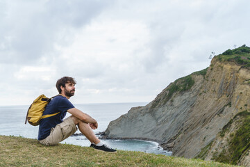 Obraz premium Adventurous caucasian man isolated sit on a bench with selective focus on the cliff. Horizontal panoramic view of man with backpack traveling in the coast. People and travel concept.