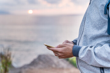 Close up with selective focus of man using smartphone. Horizontal view of man with phone with blue sea and sunset in background. Technology and people concept.