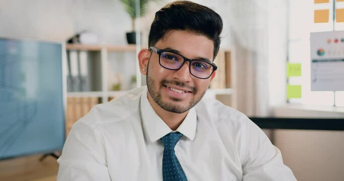 Camera view on handsome smiling successful skilled bearded engineer in glasses which sitting at workplace in design office and looking into camera with satisfied face expression