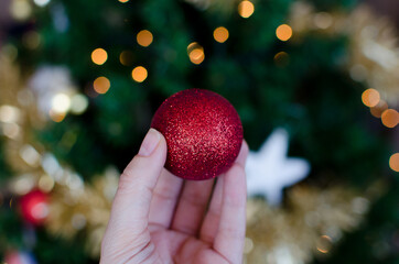 Mano humana sujetando una bola roja de purpurina con bokeh de las luces del árbol de navidad