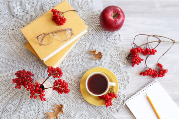 Cozy autumn days: a yellow mug with tea, viburnum branches with red berries, books, an apple, eyeglasses, a notebook on a gray napkin, top view