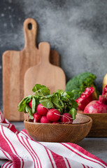 Bunch of fresh raw radish in wooden bowl on kitchen table with fresh fruit, greens, vegetables in wooden tray on grey stone background table, cooking healthy diet vegetarian food meal concept

