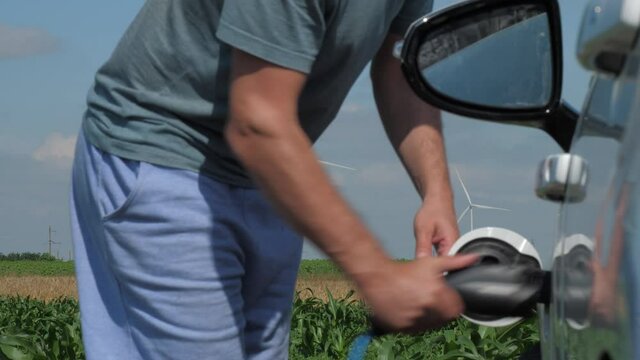 Wind turbines and e-car recharge. Man plugs off cable and gets into electric auto at station near field with wind machines on summer day closeup