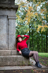 Sitting white woman with long dark hair in bright red near monument in park holding her hat