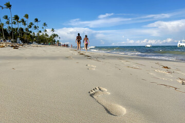 Footprints on a sand, defocused view to tropical beach with coconut palm trees and two walking women in bikini. Holiday on a paradise sea coast