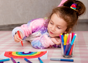 A cute girl in rainbow clothes draws a rainbow with multi-colored markers.