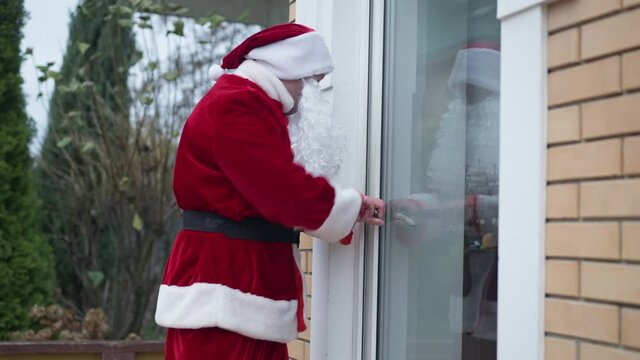 Side view burglar in red costume with white beard looking around breaking in house. Caucasian man in Santa Clause outfit opening backyard door rubbing house