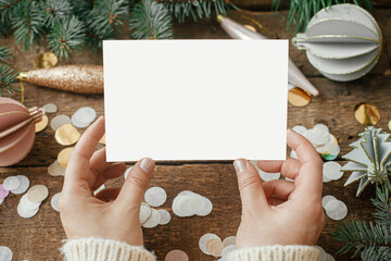 Hands holding empty greeting card and christmas ornaments, confetti, pine branches  on rustic wooden background. Christmas card mock up. Space for text. Seasons greetings. Template © sonyachny