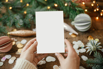 Hands holding empty greeting card and christmas ornaments, confetti, pine branches and lights on rustic wooden background. Christmas card mock up. Space for text. Seasons greetings. Template