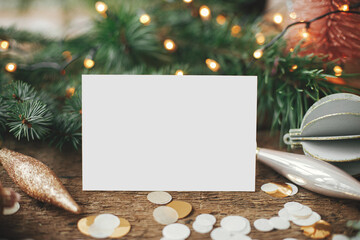 Empty greeting card and christmas ornaments, decorations, confetti, pine branches with festive lights on rustic wooden background. Christmas card mock up. Space for text. Seasons greetings template