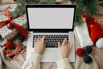 Shopping online. Hands working on laptop with blank screen on cozy bed with stylish christmas ornaments, present and santa hat. Freelance and remote work in holidays. Space for text. Top vie