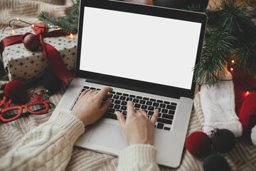Hands working on laptop with blank screen on cozy bed with stylish christmas ornaments, present and santa hat. Christmas shopping online and freelance in holidays. Space for text - obrazy, fototapety, plakaty