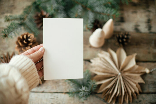 Hand holding empty greeting card on background of christmas paper stars, wooden tree, pine branches and cones on rustic wood. Christmas card mock up. Space for text. Seasons greetings template