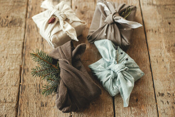 Stylish christmas gifts wrapped in fabric with fir branch and berry on rustic wooden table. Zero waste winter holidays. Furoshiki eco friendly present. Happy holidays and Merry Christmas