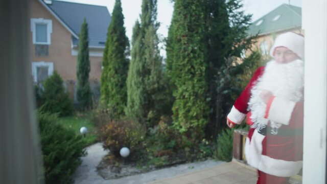 Funny Santa Clause running walking furtively with gifts bag on backyard outdoors. Shooting through glass window of cheerful Caucasian man in Father Christmas costume having fun