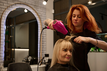 Attractive redhead woman professional hairdresser with tattooed arms in a strict black uniform combing, straightening and drying the blonde hair of beauty salon client using a pink hairdryer