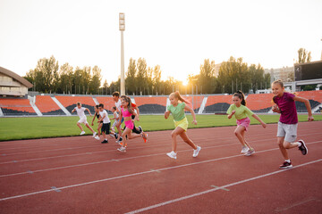 A large group of children, boys and girls, run and play sports at the stadium during sunset. A...