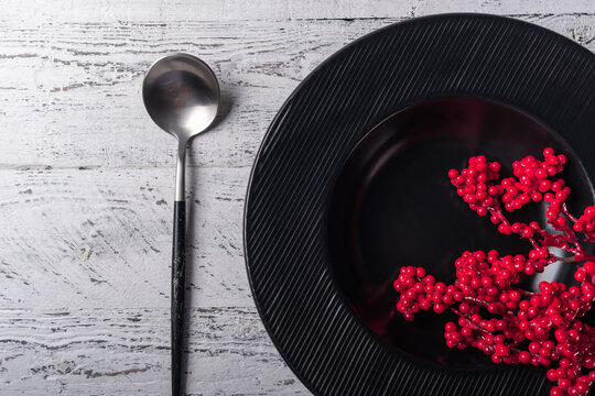 Winter table setting with a black deep plate with decorative traditional Christmas red berries, next to a tablespoon with a black shabby handle.