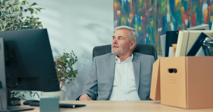 An elderly man with gray hair sits on a chair at an office desk, next to him a box of packed things, a guy in a suit looks to the side as he recalls years of working for the company he is retiring