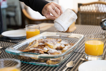Hand of woman adding powdered sugar on french toasts tray by orange juice glasses on breakfast table