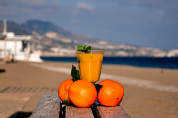 A glass with orange juice and a sprig of mint surrounded by three oranges is on a wooden table against a blurred background of the beach, sea, mountains and resort town in clear weather.