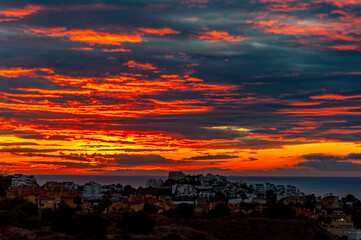 View of the coastal city against the backdrop of the sea and sky with clouds tinted red from the sun during the morning twilight before sunrise;