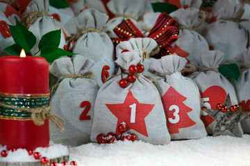 Close-up advent calendar diy sacks of gray canvas tied with twine,blurred candle on side,Christmas decor,red berries,wooden star,Poinsettia leaves and snow in foreground. Celebrating and gifts concept