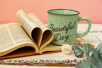 A green cup of tea stands on a book. Pink background. Eucalyptus flowers. The pages of the book are wrapped in the shape of a heart