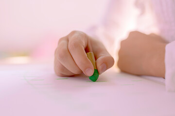 Hand of a girl drawing with a green wax on a paper in a room with soft lighting with pink tones