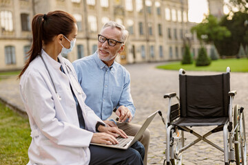 Cheerful mature recovering patient with wheelchair having conversation with young female doctor working on a laptop in the park near clinic