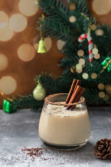 Christmas eggnog with cinnamon sticks on a background of fir branches decorated with toys, blurred bokeh. Glass of a festive winter drink on a concrete background with copy space. 