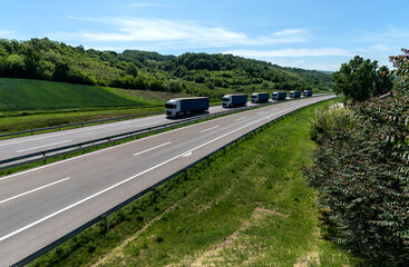 A convoy of trucks heading down the highway between meadows and dense forests. White clouds are covering the sky. Caravan or convoy of trucks in line on a country highway.