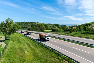 A convoy of trucks heading down the highway between meadows and dense forests. White clouds are covering the sky. Caravan or convoy of trucks in line on a country highway.	
