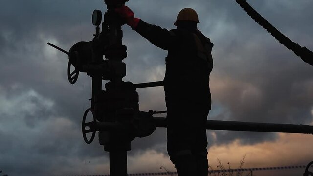 An employee in overalls and a helmet is carrying out repairs and maintenance of an oil well. Silhouette on the background of the evening sky