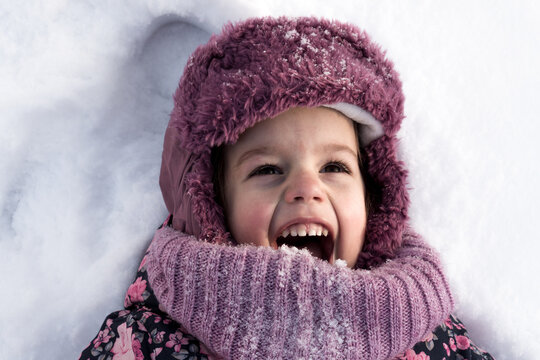 Winter, family, childhood concepts - close-up portrait authentic little preschool girl in pink clothes smile laugh shout with open mouth laying on snow in frosty weather day outdoors. Funny kid face
