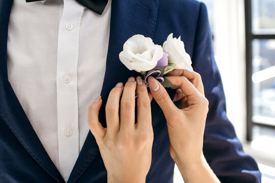 On the wedding day, the bride helps the groom to put on a boutonniere of white roses on his jacket. Close-up - the bride's hands and the groom's classic blue suit. Caring bride helps her groom