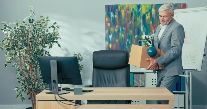 Ambitious older man with gray hair wearing a shirt stands in front of desk in corporate office holding box of packed items in hands comes to corporation to be promoted to new job unpacking accessories