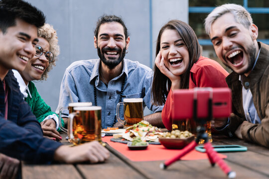 Group of multiracial friends enjoying food and drinking beer while making online video call with mobile phone at brewery bar to celebrate birthday - Focus on bearded man face