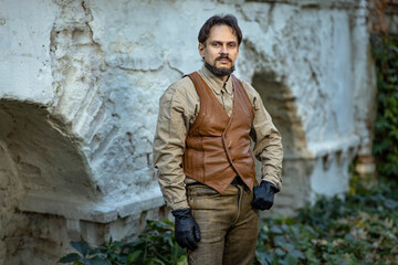 Fototapeta na wymiar A 19th century man in a leather vest and black gloves against the background of the wall of an old fortress