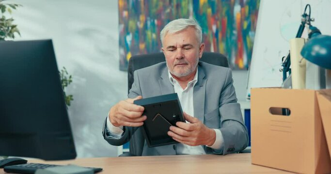 New employee older man with gray hair wearing shirt sits at desk in office holding box of packed items next to him, removes picture frame places on counter next to computer prepares workstation