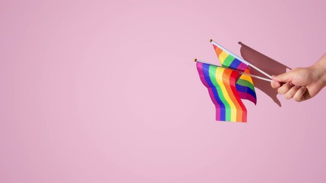 hairy hand of a man holding rainbow flag above a pink background. hard shadows of the sun, vertical orientation, copy space