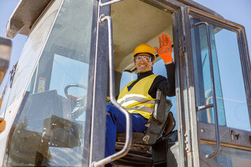 Cheerful male worker in safety helmet waving hand and smiling while sitting in driver cabin of...
