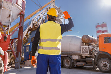 Back view of man engineer wearing work vest and safety helmet while looking at industrial equipment and truck at production plant