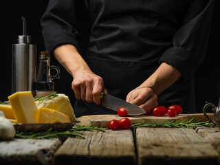 The chef is cutting tomatoes on a cutting board. Ingredients for making salad, sauce, pizza. Wooden texture, black background. Home cooking, hotel, restaurant, cafe, culinary blog.
