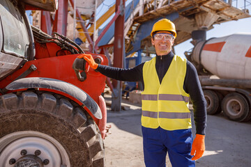 Cheerful man in work vest and safety helmet looking at camera and smiling while placing hand on truck