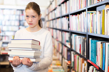 Sad tired preteen schoolgirl standing in library with pile of textbooks in hands.