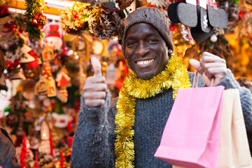 Portrait of laughing African-American guy holding bags after shopping on outdoor Christmas market.