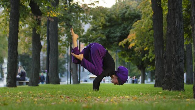 Female muslim teacher in hijab practice yoga flow power asana in city park on grass workout for healthy body strong spine pose athletic legs movement in air. Woman girl feel energy training bakasana
